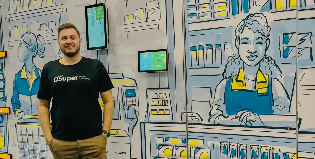 Homem sorrindo ao lado de um painel ilustrado que representa um ambiente de supermercado, com telas digitais exibindo informações no fundo. Ele veste uma camiseta preta com o logotipo OSuper Digital Commerce